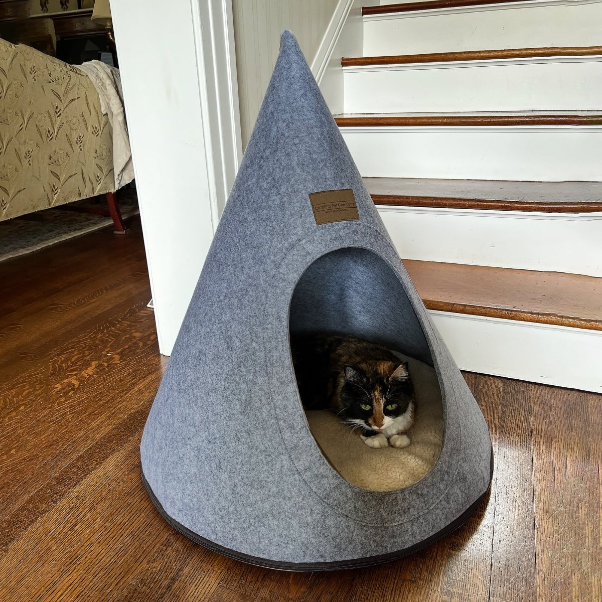 Gray pet bed shaped like a cone with a cat peeking out, placed on a wooden floor.