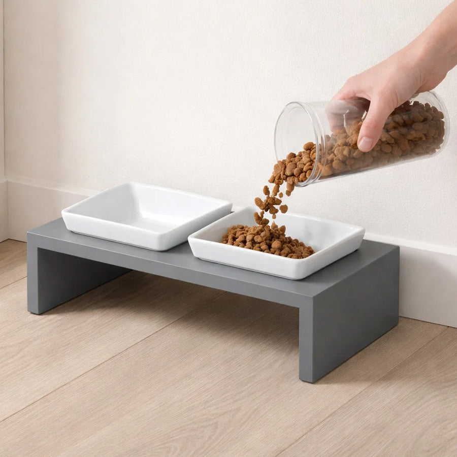 A two-bowl elevated pet feeder stand in gray with white square bowls, shown on a light wood floor against a beige wall. A human hand is pouring kibble from a clear container into one of the ceramic white bowls.