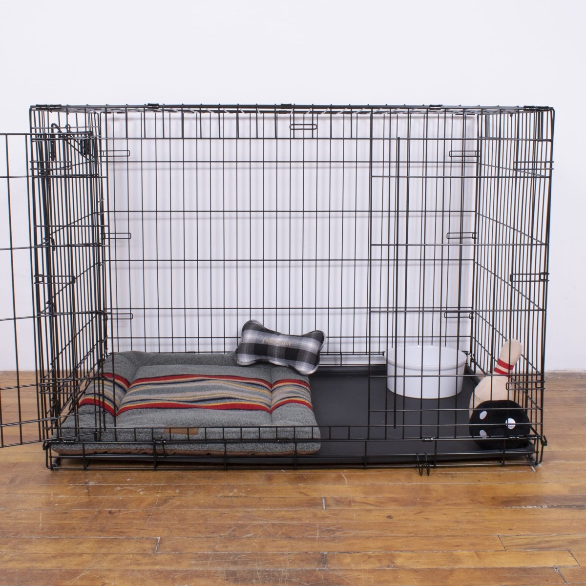 Dog crate with mat, bedding, toy, and water bowl on a wooden floor.