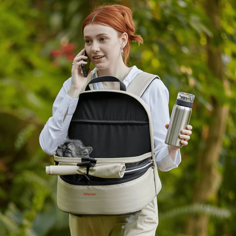 Person with red hair using a phone, carrying an ibiyaya pet backpack front facing with a cat, and holding a thermos outdoors.